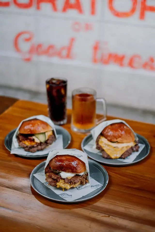 Three smash burgers with fries, a cola and a beer, in front of a Curators of Good Times wall